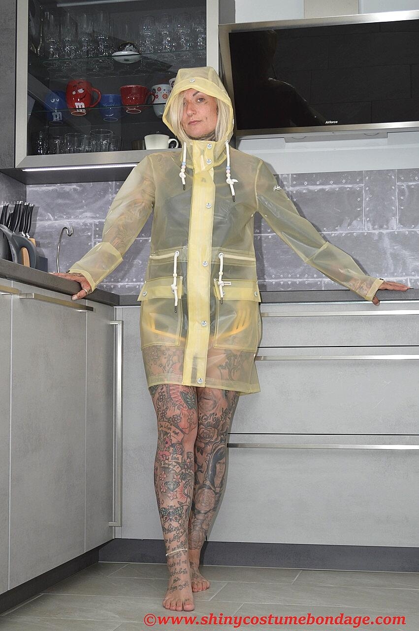 Tattooed blonde Miss Francine models barefoot in a raincoat over rubber wear